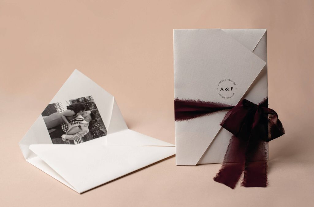 Convites de casamento sob uma tira de tecido bordô sob fundo rosa.