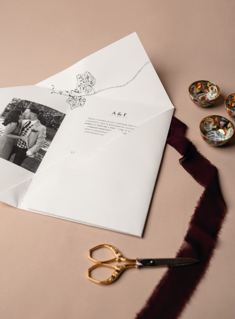 Convites de casamento sob uma tira de tecido bordô, com uma tesoura dourada, sob fundo rosa.