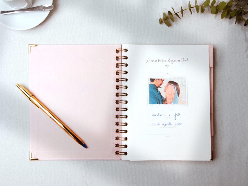 Agenda da noiva cor de roza com palavras em foil dourado e fita branca e uma caneta dourada