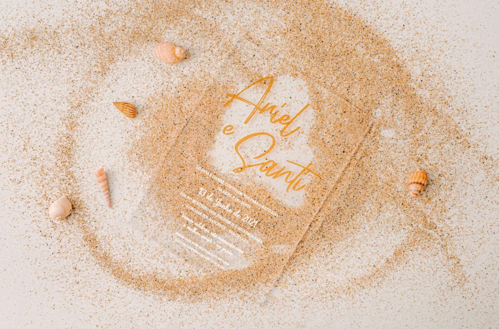Convite de batizado de acrílico com impressão a branco e foil dourado pousado em areia.