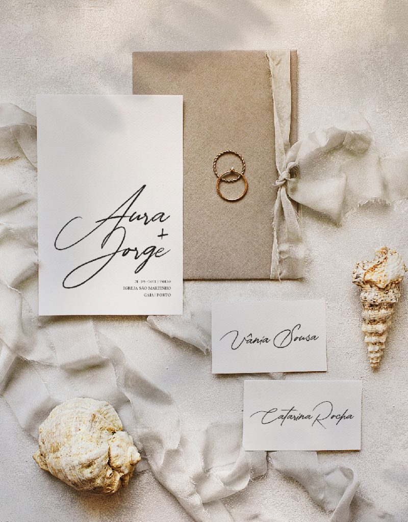 Convite de casamento de estilo tipográfico com um laço de linho bege, com um envelope cinza pousado numa bandeja douradaConvite de casamento de estilo tipográfico com um laço de linho bege, com um envelope cinza pusado numa bandeja dourada