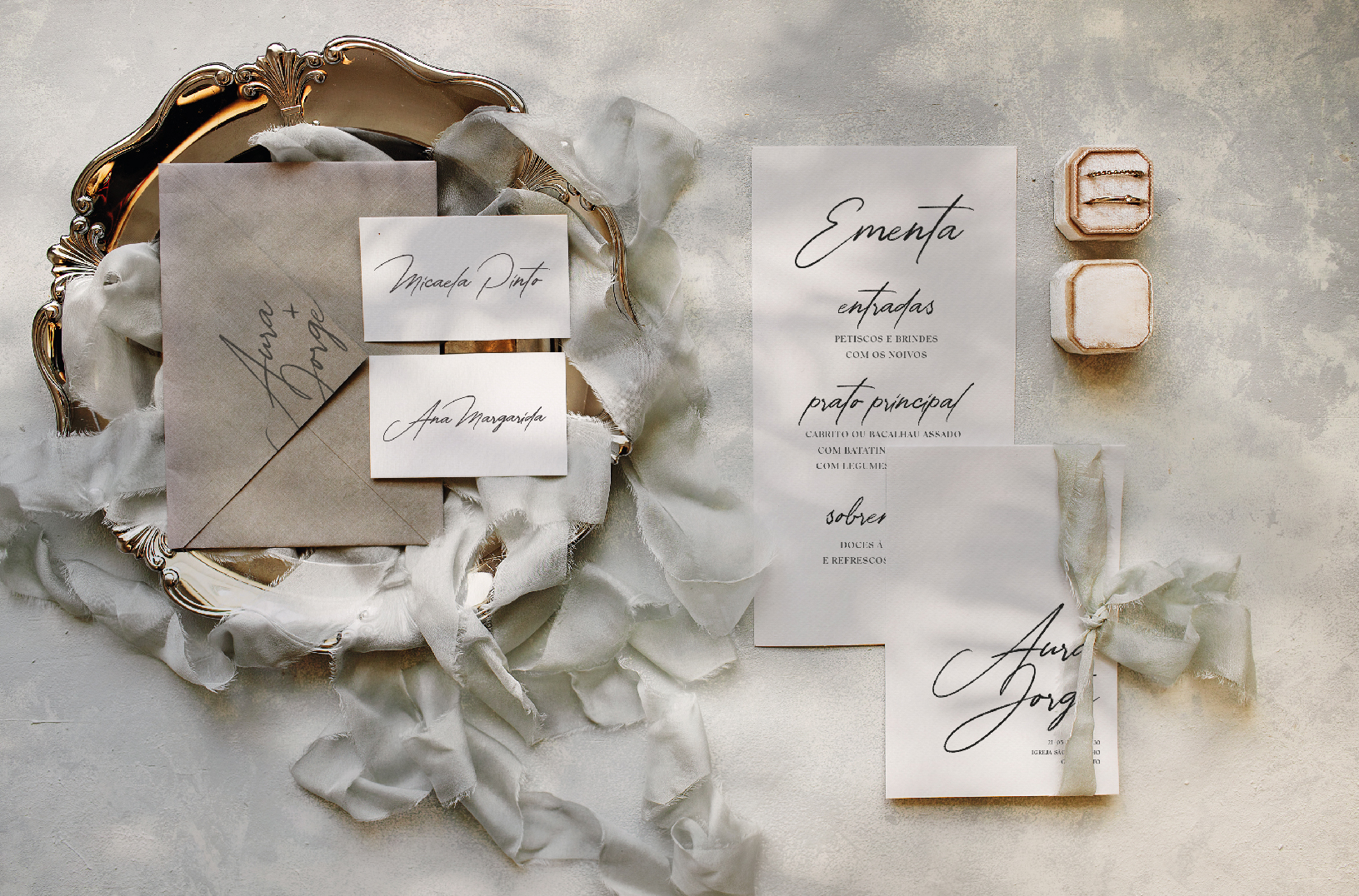 Convite de casamento de estilo tipográfico com um laço de linho bege, com um envelope cinza pousado numa bandeja dourada.