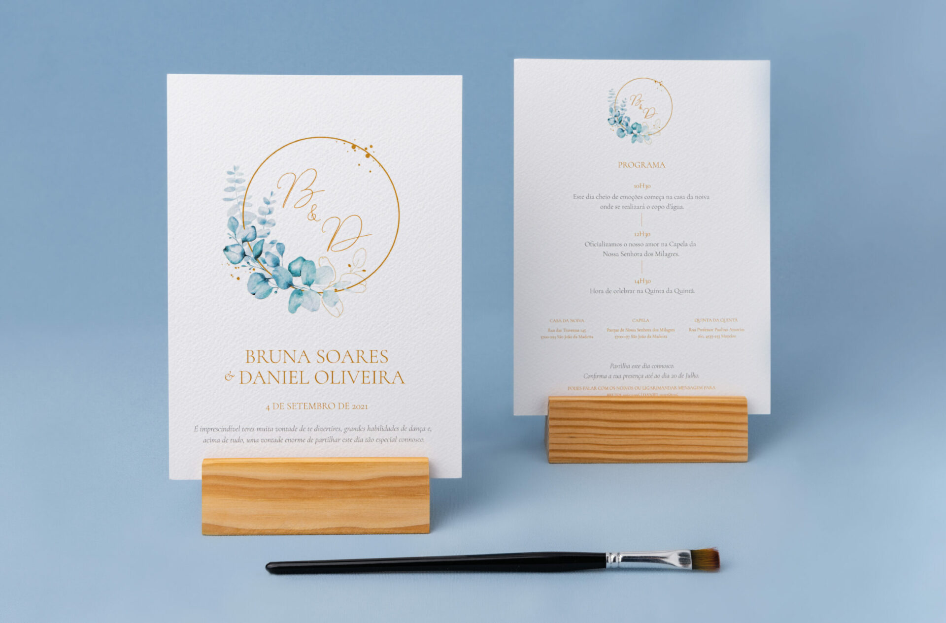 Convite de Casamento branco de estilo Aguarela com tipografia dourada e ilustração em aguarela pousado numa base de madeira e fundo azul
