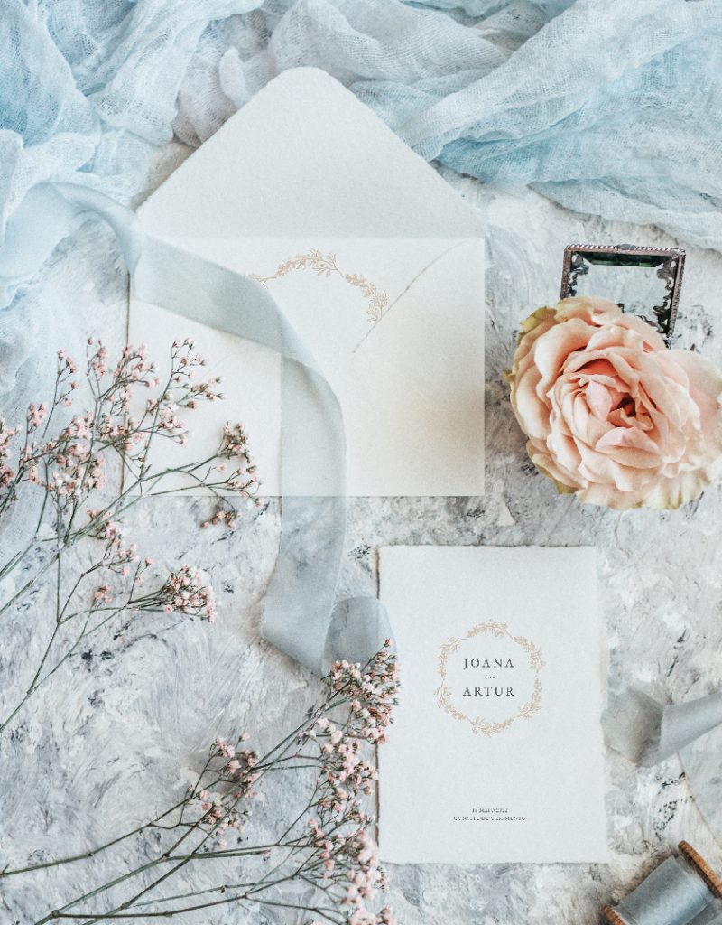 Convite de casamento branco de estilo tipográfico com acabamento rasgado, com um envelope branco com impressão interior com fitas rosa e azul e flores rosa